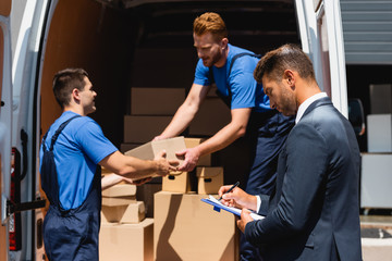 Obraz premium Selective focus of businessman writing on clipboard while movers holding carton box in truck outdoors