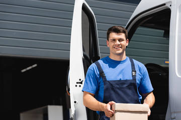 Young loader holding carton box and looking at camera near truck outdoors