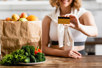 partial view of woman holding credit card near paper bag and plate with vegetables on table