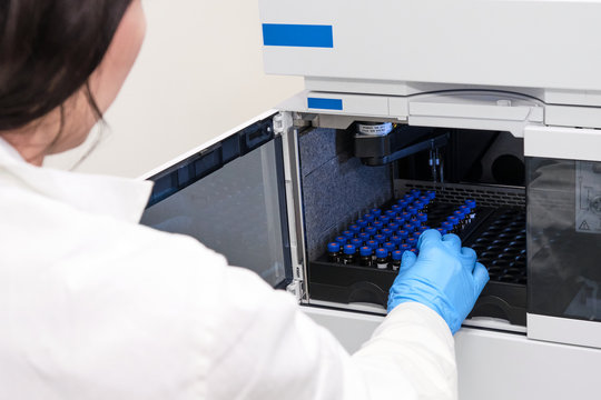 Scientist in a white lab coat putting vial with a sample into autosampler of HPLC system. High performance liquid chromatography at chemical laboratory. Developing of pharmaceuticals or vaccine