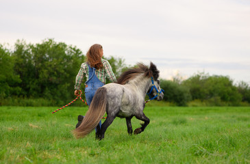 The young Caucasian woman and her gray pony are running on the grass in rural.