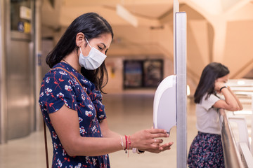 Young mother on new normal using the alcohol antiseptic gel. Washing hands using sanitizer dispenser while her daughter waits on the background.