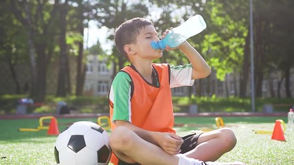 Handsome tired sporty 13-aged footballer sitting on the special grass covering of the outdoors sport ground and dringking water after training on sunny day