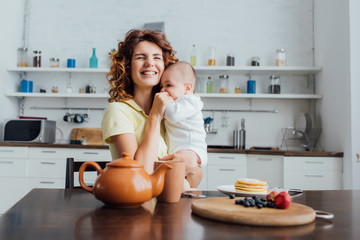 selective focus of young mother holding infant boy in rompers near table served with breakfast