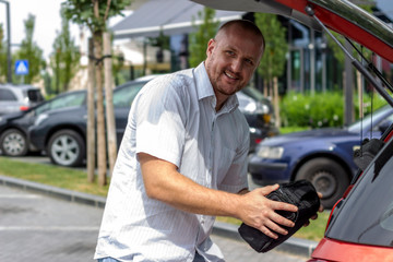 Photo of man putting bags in the car trunk. Businessman is unpacking the car trunk.Man packing luggage in car ready for road trip.Young Caucasian man loading the trunk of the car parked on the parking