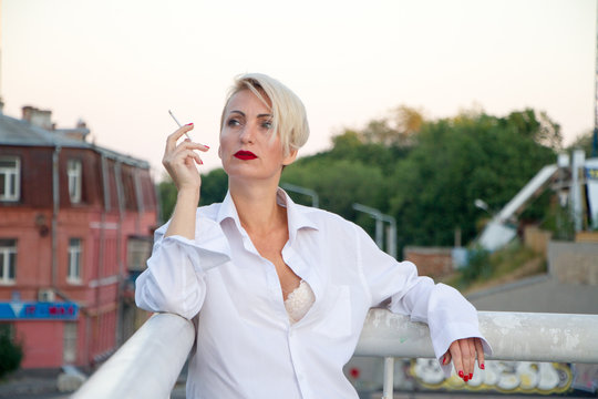 Beautiful mature blonde woman in white shirt smokes white cigarette outdoors in city at summer