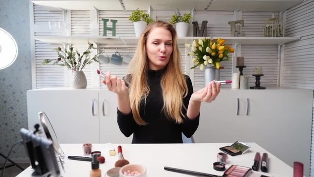 Vlogger female applies lipstick on lips. Beauty blogger woman filming daily makeup routine tutorial at camera on tripod. Influencer blonde lady live streaming cosmetics product comparison in studio