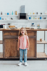 full lengtn view of girl standing near kitchen table with ingredients and cooking utensils