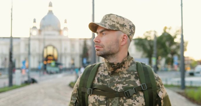Caucasian young brave male soldier in uniform, hat and with backpack walking outdoor. Confident militarian officer strolling the street in military clothes. Going to army or coming back.