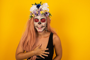 Joyful European student keeps hands crossed, laughs at good joke, wears casual clothes and round spectacles, standing indoors. Happy young woman with long hair poses against gray wall.