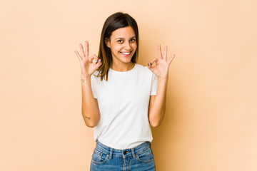 Young woman isolated on beige background cheerful and confident showing ok gesture.