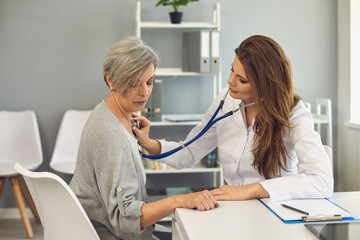 Fototapeta premium Female doctor listens with stethoscope to patient. Senior man sitting in clinic office.