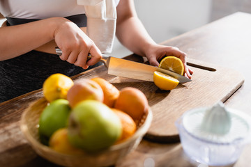 cropped view of young woman cutting fresh lemon on chopping board near fruits and juicer