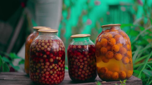 Jars with kompot on table. Sealed glass jars with kompot placed on table near flowers and fruits on summer day in garden
