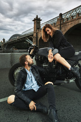 Fototapeta na wymiar Romantic date on motorbike. Young woman sits on a motorcycle and looks at the man who sits on the ground and holds her hand. Couple in love with sunset under the bridge in the city