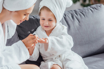 selective focus of young mother making manicure to daughter in white bathrobe and towel on head