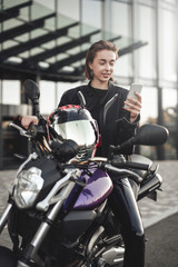 Fototapeta na wymiar Funny girl in a motorcycle jacket sits on a purple motorbike and looks at her phone