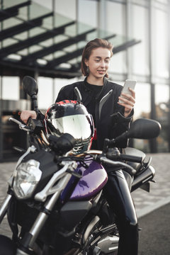 Funny girl in a motorcycle jacket sits on a purple motorbike and looks at her phone