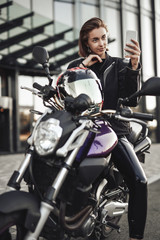 Girl in a motorcycle jacket sits on a purple motorbike and looks at her phone