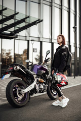Beautiful young brunette girl in a black motorcycle jacket stands near her purple sport motorbike