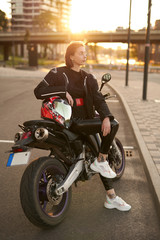 Very beautiful girl in a black motorcycle jacket sits with red and black motorcycle helmet on a black sport motorbike