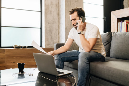 Mature man sitting on couch, using laptop, talking on the phone, holding papers