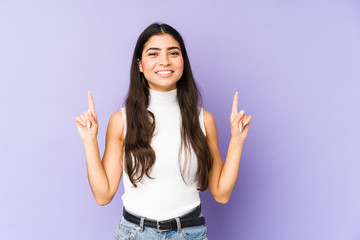 Young indian woman isolated on purple background indicates with both fore fingers up showing a...