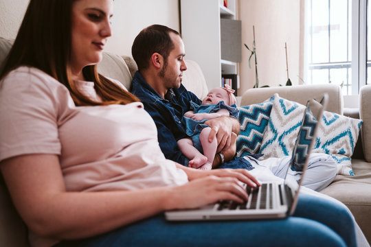 Father carrying sleeping daughter while mother using laptop on sofa