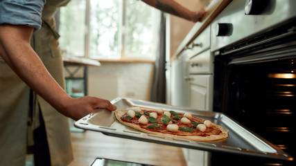 Cropped shot of man, professional cook making pizza at home. Man in apron putting raw pizza in modern oven for baking.