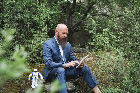 Bald businessman using digital tablet while sitting with robot amidst trees in forest