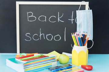 blackboard with inscription back to school on it, medical mask hang on the corner. School supplies lie on the blue table near. Back to school concept. After pandemic restrictions.