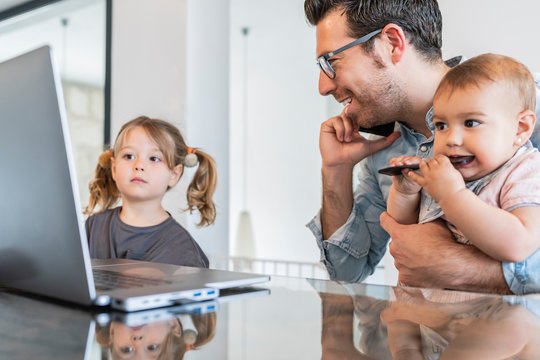 Man holding daughter talking over smart phone while girl looking at laptop on table