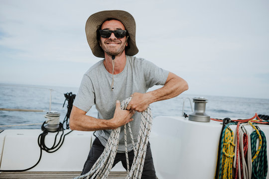 Smiling sailor wearing sunglasses and hat holding ropes in sailboat