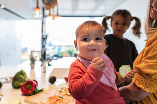 Close-up of cute baby girl eating zucchini while mother and daughter working in background