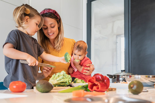 Mother holding baby girl while guiding daughter in cutting vegetables on kitchen island