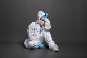 Tired female doctor in a protective suit and mask sitting on the floor on a gray background. Emergency medicine and ambulance during COVID-19.