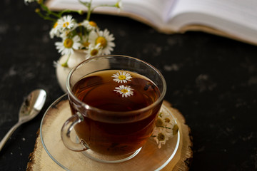 English tea with chamomile stands on a wooden frame and a teaspoon and an open book for reading lies on a black background. Nearby is a white vase with summer chamomile flowers