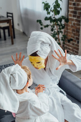 Fototapeta na wymiar high angle view of mother in face mask showing scaring gesture while sitting with daughter in bathrobes and towels on heads