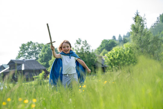 Playful boy wearing cape holding toy sword while running on grassy land against clear sky