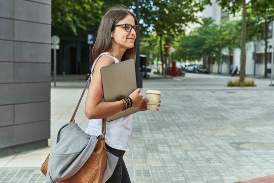 Thoughtful businesswoman holding laptop and disposable glass while walking on city street
