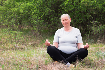 Naklejka na ściany i meble Portrait of a senior woman, wearing a white T-shirt and black pants, meditates in nature. Meditation, yoga and relaxation concept.