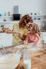 selective focus of mother holding rolling pin and daughter scattering flour while standing face to face at kitchen table