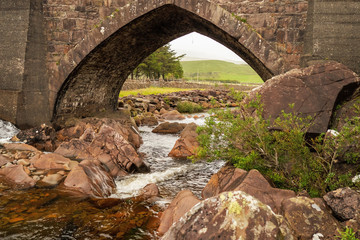 Fototapeta na wymiar Small river with brown color rocks flows under small old bridge. West of Ireland. Connemara.