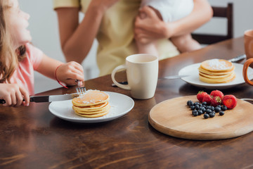 cropped view of mother holding infant son near daughter eating delicious pancakes, selective focus