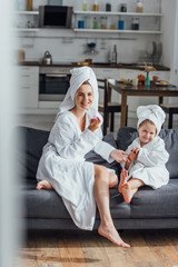 selective focus of young woman holding nail file while sitting on sofa with daughter in terry bathrobes and towels on heads