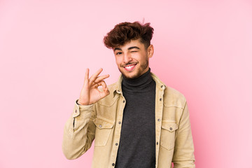 Young arabian man posing in a background isolated winks an eye and holds an okay gesture with hand.