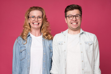 Funny caucasian woman and man smiling happily while looking at camera, standing over pink background