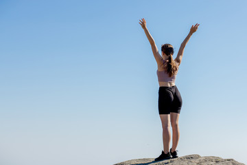 Obraz premium The young girl at the top of the mountain raised her hands up on blue sky background. The woman climbed to the top and enjoyed her success. Back view