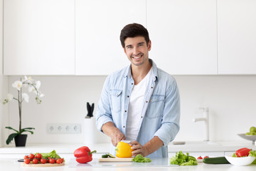 Young man cutting vegetables for salad at table in modern light kitchen.