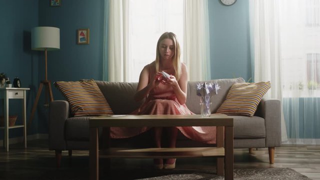 A young girl in a pink dress sits on the couch, twists a white sheet in his hands and makes it origami on a coffee table